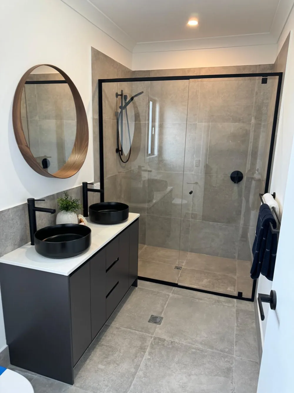 Modern grey-tiled bathroom with black double basin vanity, round timber mirror and black-framed shower